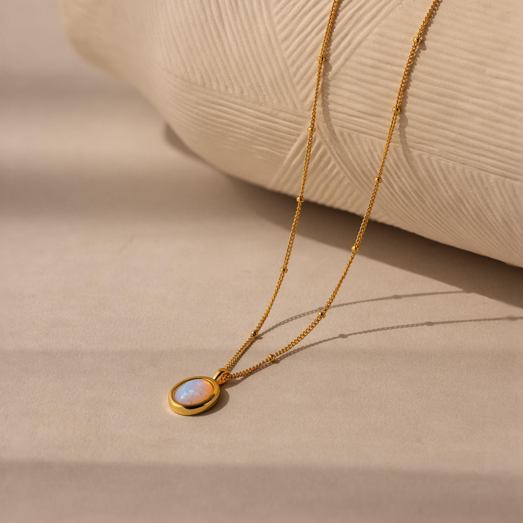 The Opal Pendant Necklace displays an oval opal gemstone on a dainty satellite chain, shown resting on a beige textured surface.