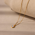The Opal Pendant Necklace displays an oval opal gemstone on a dainty satellite chain, shown resting on a beige textured surface.