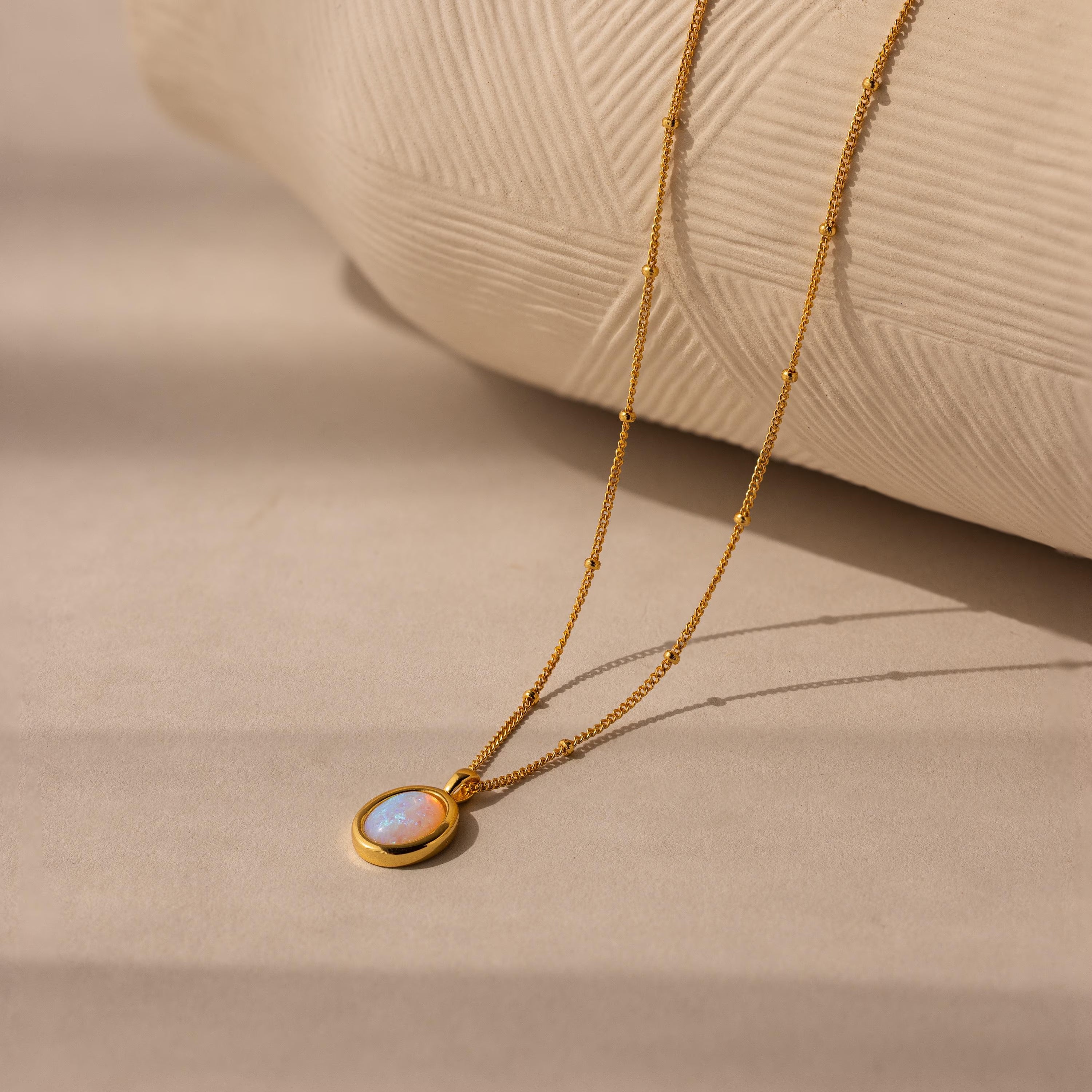 The Opal Pendant Necklace displays an oval opal gemstone on a dainty satellite chain, shown resting on a beige textured surface.