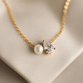 The Pearl Diamond Necklace features a gold chain adorned with a pearl and teardrop diamond, beautifully displayed on a soft beige surface—a timeless Toi et Moi piece.