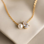 The Pearl Diamond Necklace features a gold chain adorned with a pearl and teardrop diamond, beautifully displayed on a soft beige surface—a timeless Toi et Moi piece.