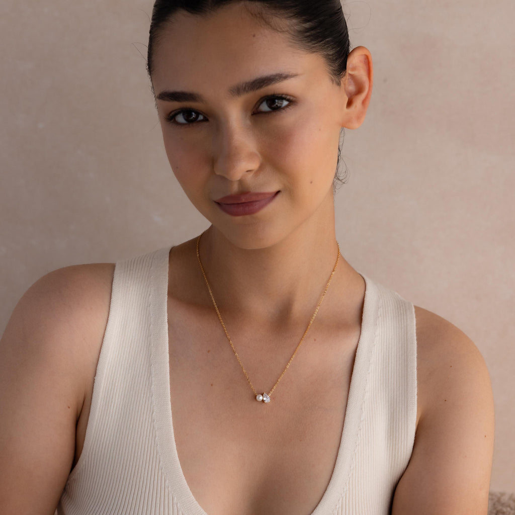 A woman with dark hair in a bun wears a cream sleeveless top and the Pearl Diamond Necklace, featuring a delicate design with a small pendant.