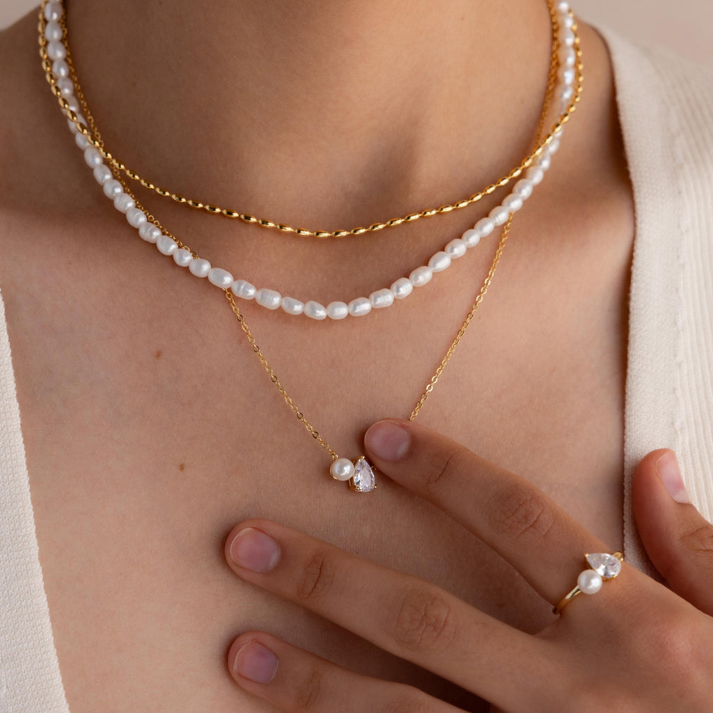 Close-up of a woman wearing layered gold and Pearl Diamond Necklaces (featuring the Pearl Diamond Necklace), a teardrop diamond ring, and a cream top.