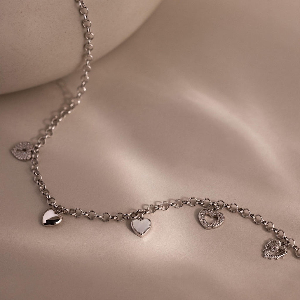Silver heart charm necklace with five unique heart pendants displayed on a soft beige background.