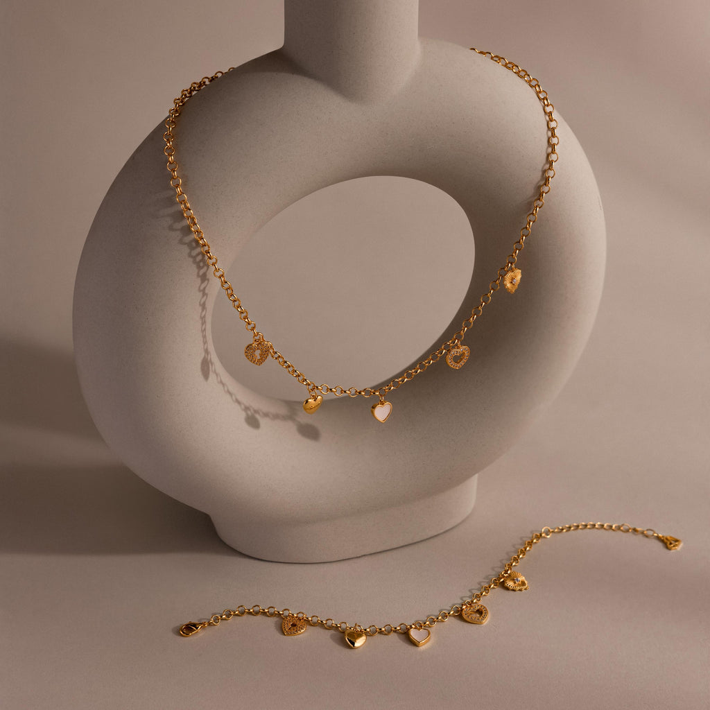 Gold heart charm necklace and matching bracelet displayed on a ceramic sculpture, featuring multi-style heart pendants and chunky rolo chain.