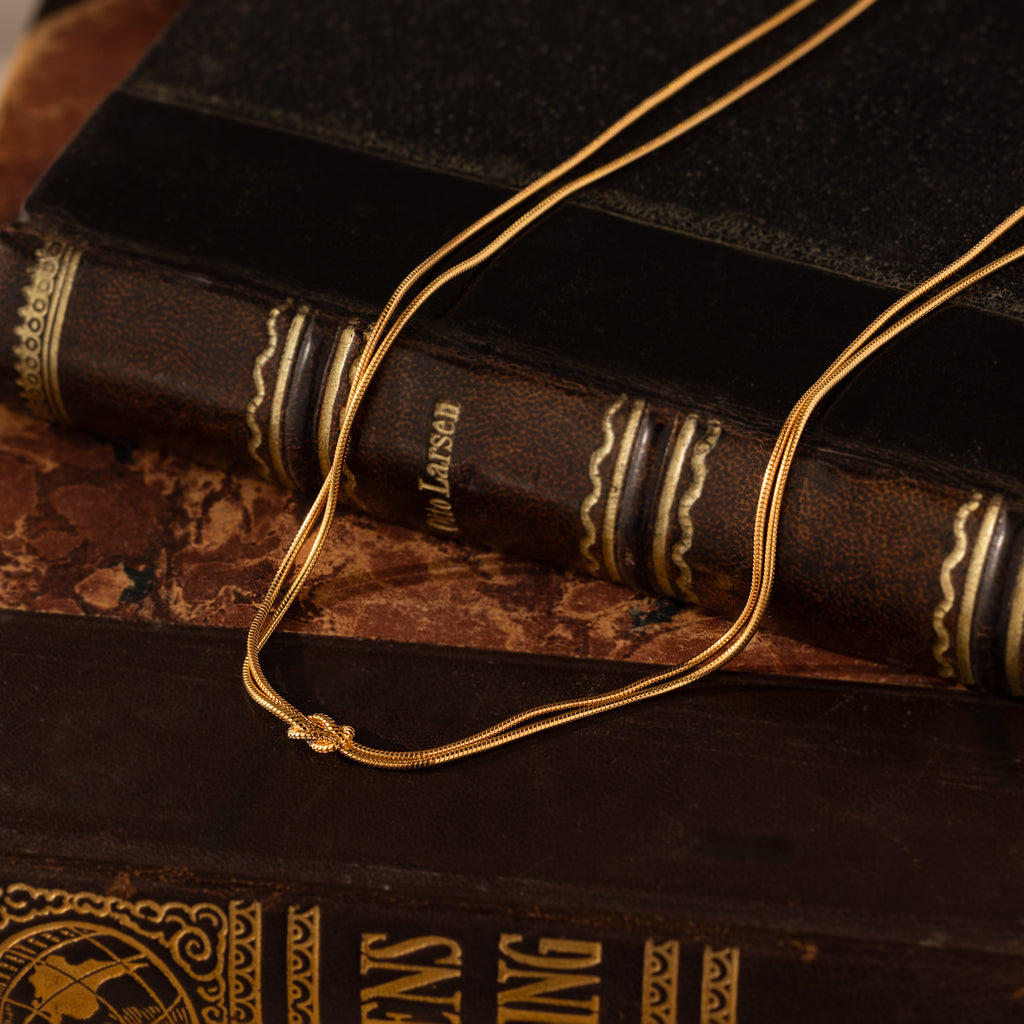 A Love Knot Chain Necklace in 18K Gold rests atop two vintage books with ornate spines, symbolizing meaningful bonds that transcend generations.