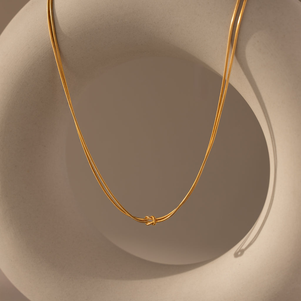The Love Knot Chain Necklace in 18K Gold, symbolizing meaningful bonds, is draped over a smooth, beige abstract sculpture.