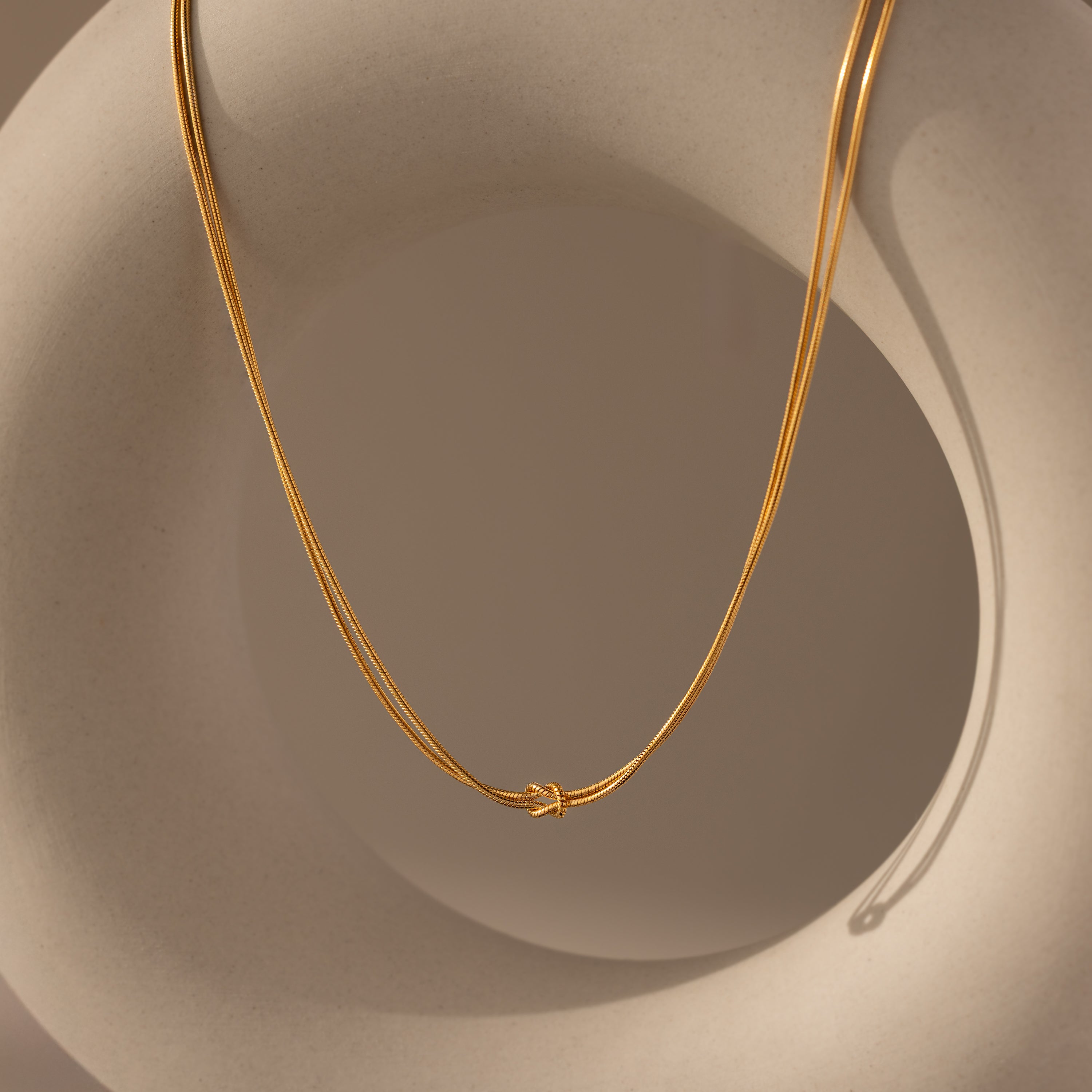 The Love Knot Chain Necklace in 18K Gold, symbolizing meaningful bonds, is draped over a smooth, beige abstract sculpture.