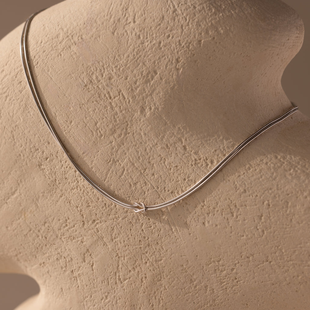 The Love Knot Chain Necklace in White Gold is displayed on a textured beige mannequin bust, symbolizing meaningful bonds.