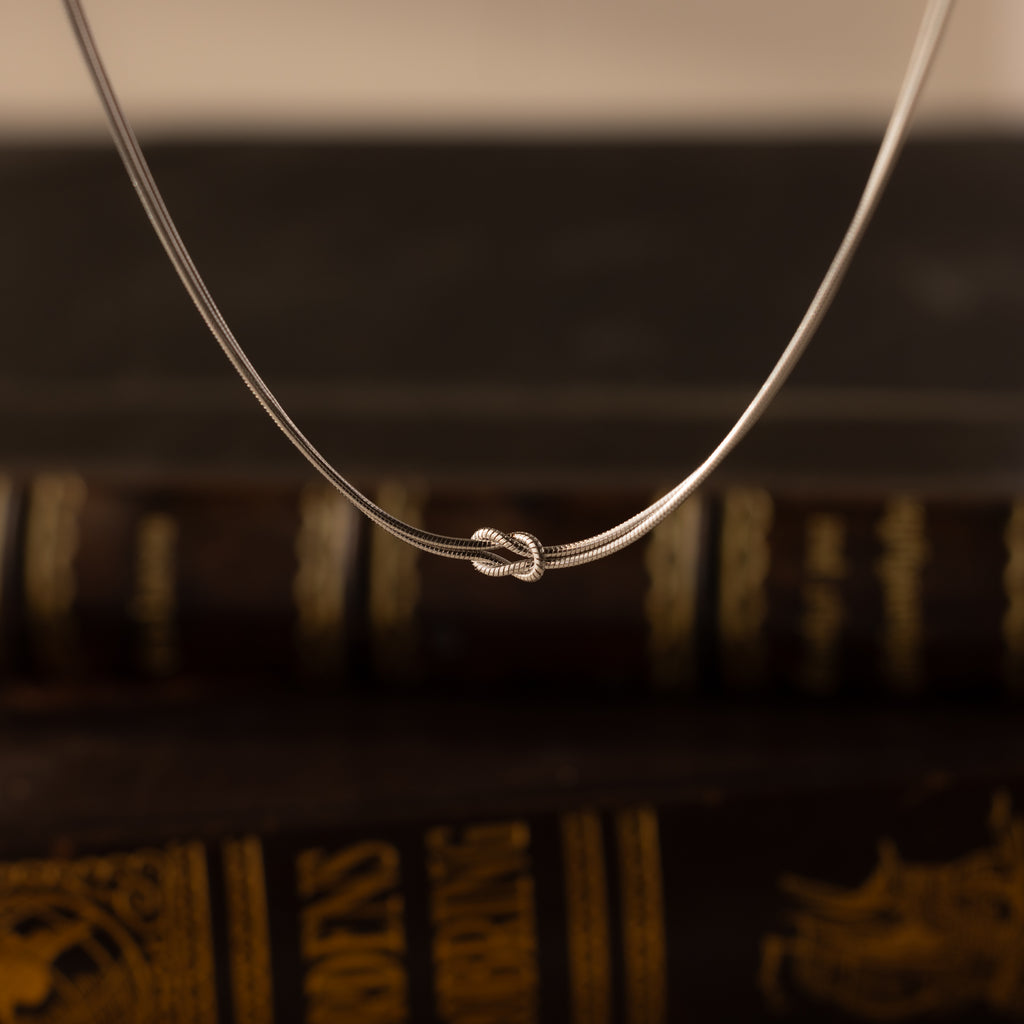 The Love Knot Chain Necklace in White Gold, symbolizing meaningful bonds, is displayed against softly blurred vintage books.