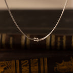 The Love Knot Chain Necklace in White Gold, symbolizing meaningful bonds, is displayed against softly blurred vintage books.