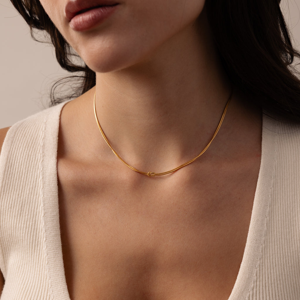 A woman wears the Love Knot Chain Necklace in 18K Gold, a symbol of meaningful bonds, paired with a cream V-neck top; she is shown from shoulders to lips.
