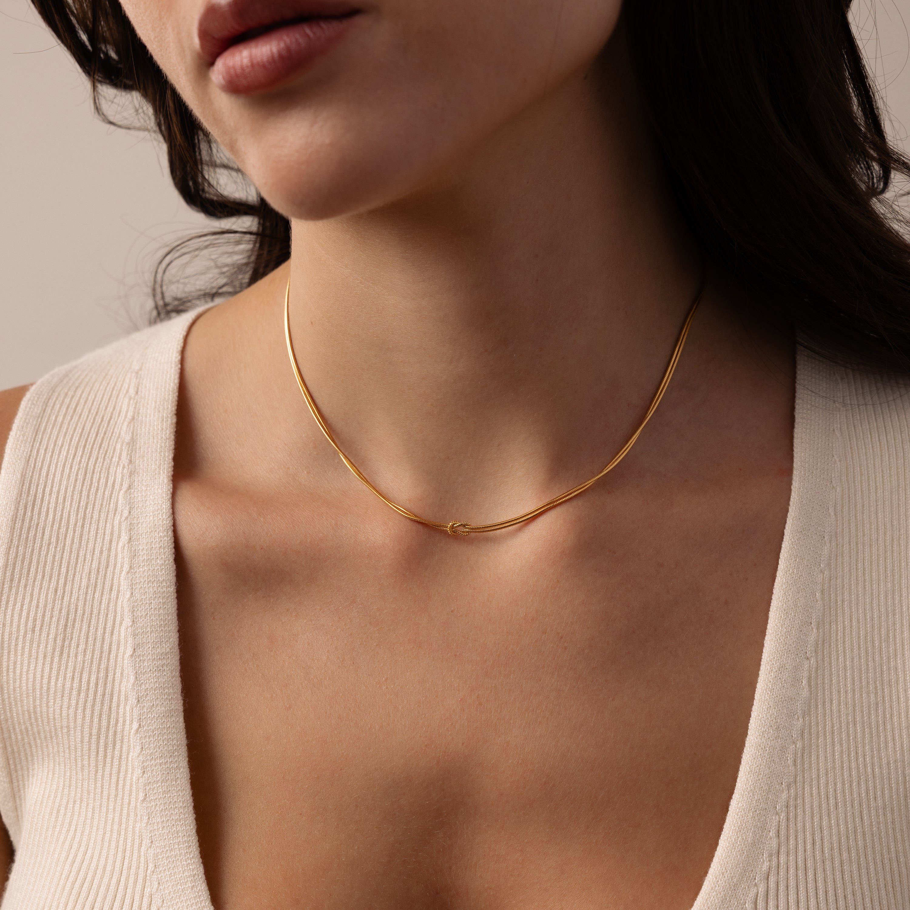 A woman wears the Love Knot Chain Necklace in 18K Gold, a symbol of meaningful bonds, paired with a cream V-neck top; she is shown from shoulders to lips.