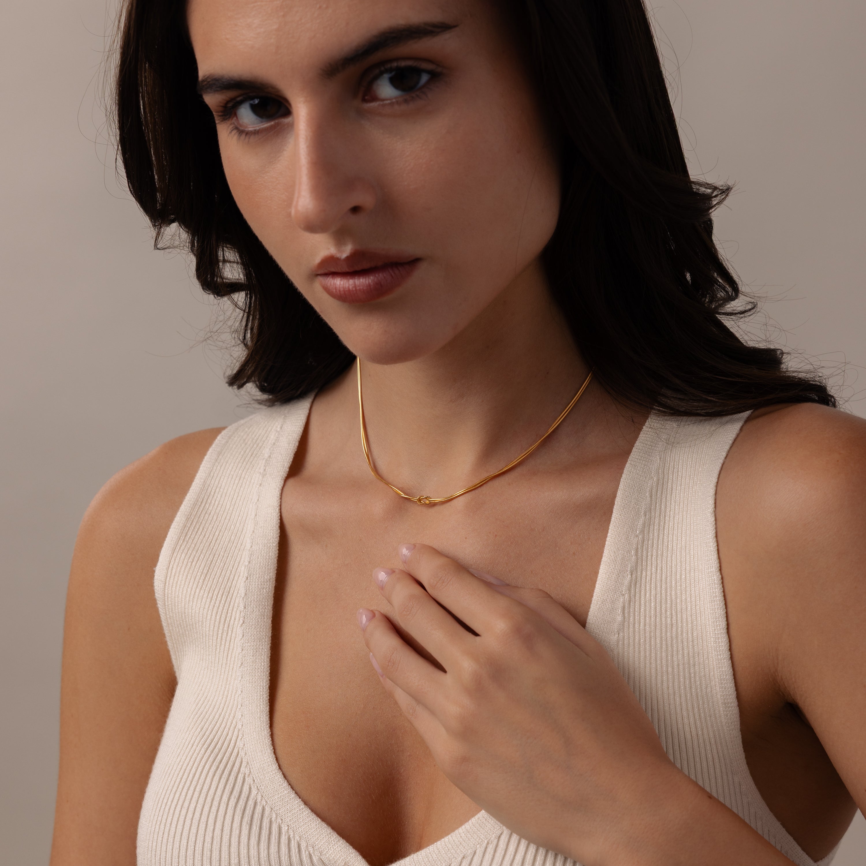 A woman in a cream top wears the Love Knot Chain Necklace in 18K Gold, gently touching it as she looks at the camera—a graceful reminder of cherished connections.