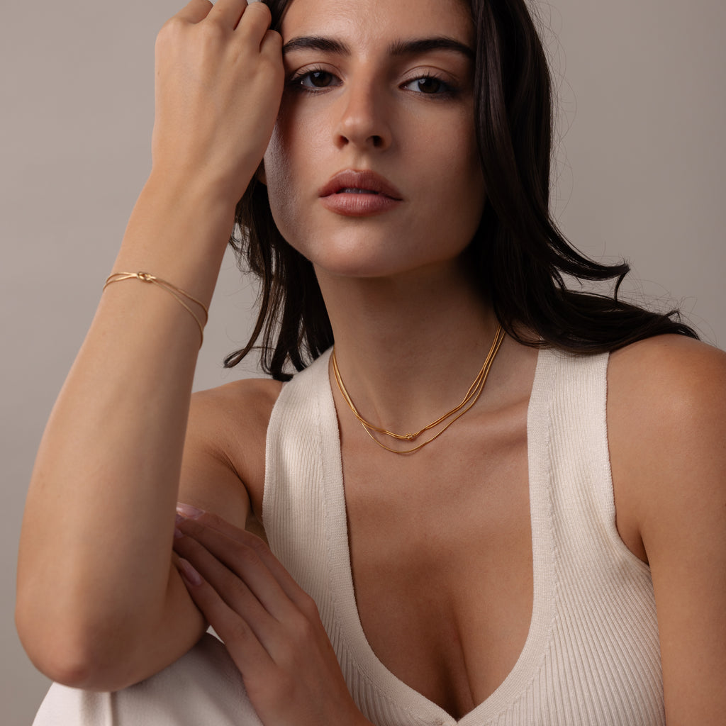 Woman in a cream sleeveless top wears gold jewelry, including the Love Knot Chain Necklace in 18K Gold, symbolizing meaningful bonds, and poses with one hand near her face against a neutral background.