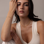 Woman in a cream sleeveless top wears gold jewelry, including the Love Knot Chain Necklace in 18K Gold, symbolizing meaningful bonds, and poses with one hand near her face against a neutral background.