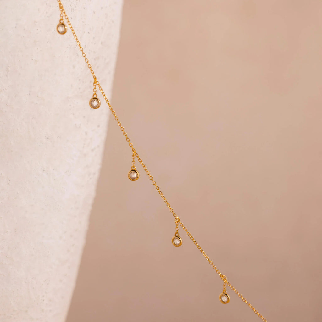 18k gold chain with round bezel-set diamond  crystals spaced delicately along its length against a soft neutral background.