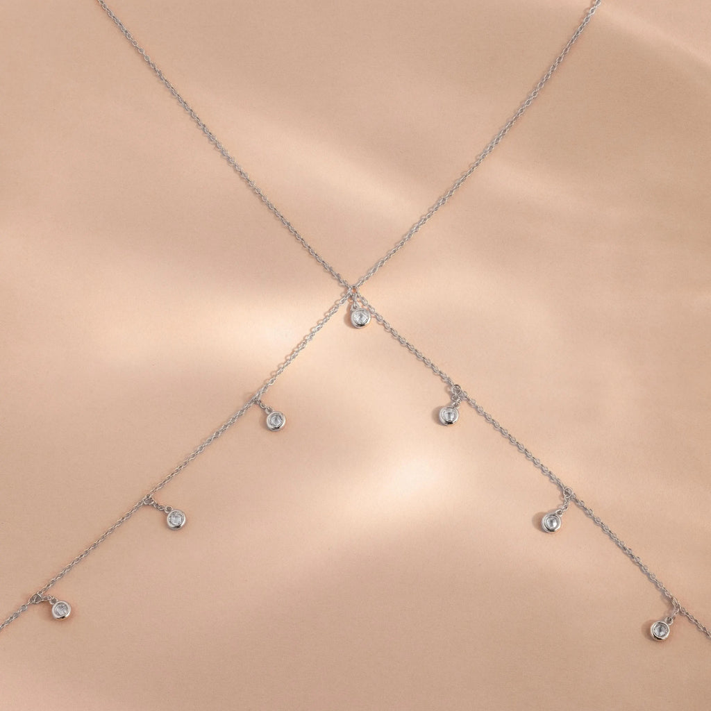 Close-up of the sterling silver Diamond Station Body Chain laid out in an X-shape on a soft blush satin backdrop.
