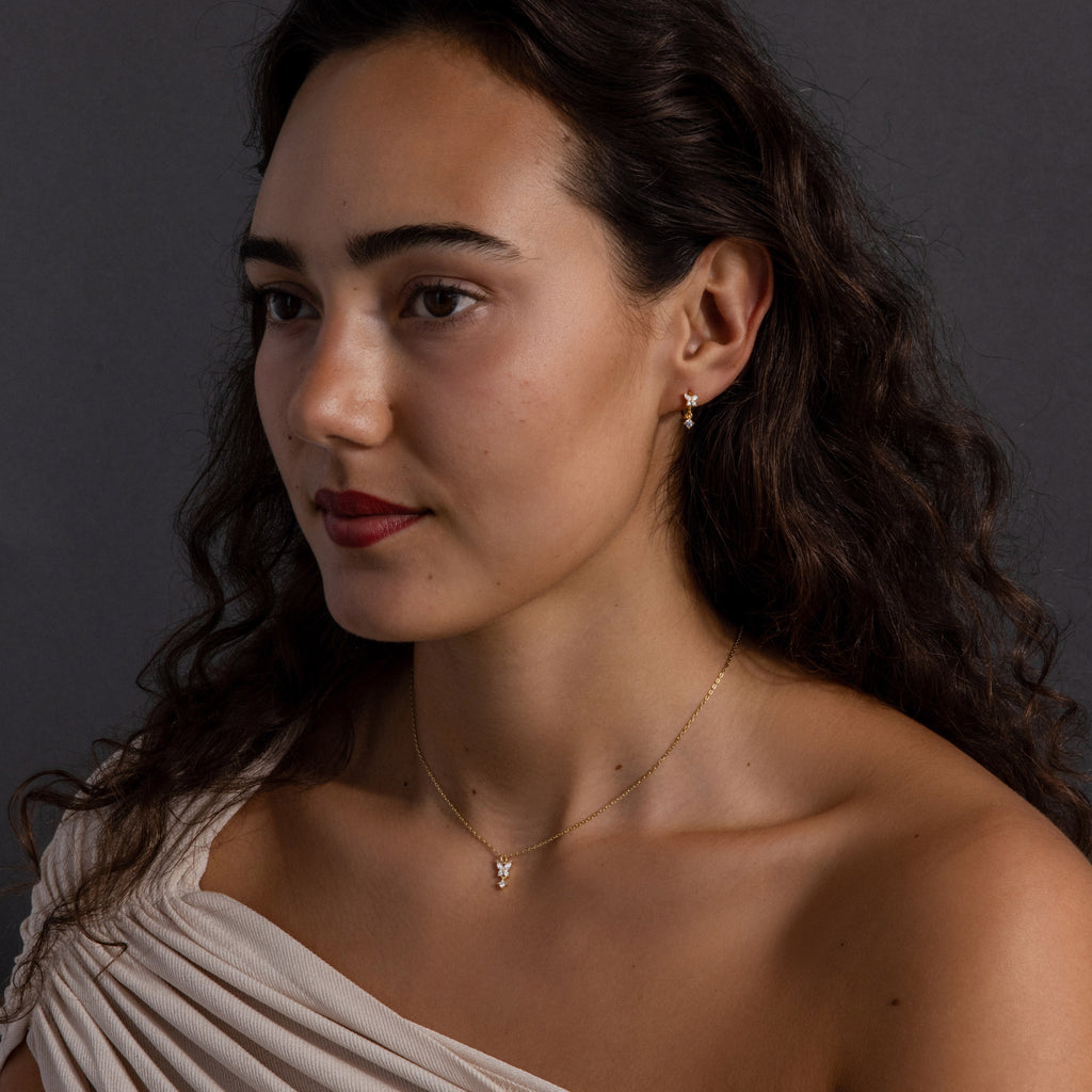 Woman with long curly hair wearing a white top, gold earrings, and the Diamond Butterfly Drop Jewelry Set in 18K Gold, posing against a dark background.