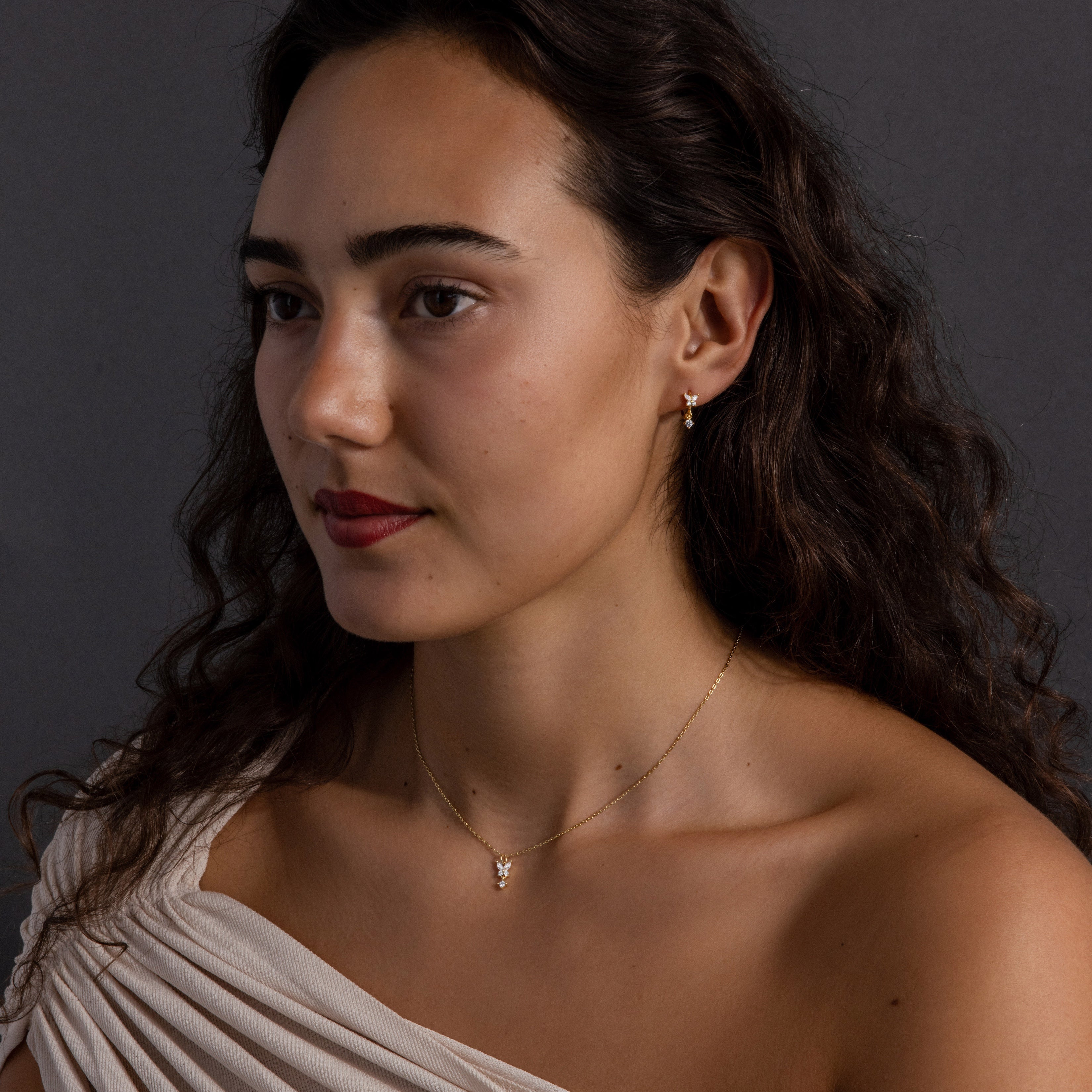 Woman with long curly hair wearing a white top, gold earrings, and the Diamond Butterfly Drop Jewelry Set in 18K Gold, posing against a dark background.