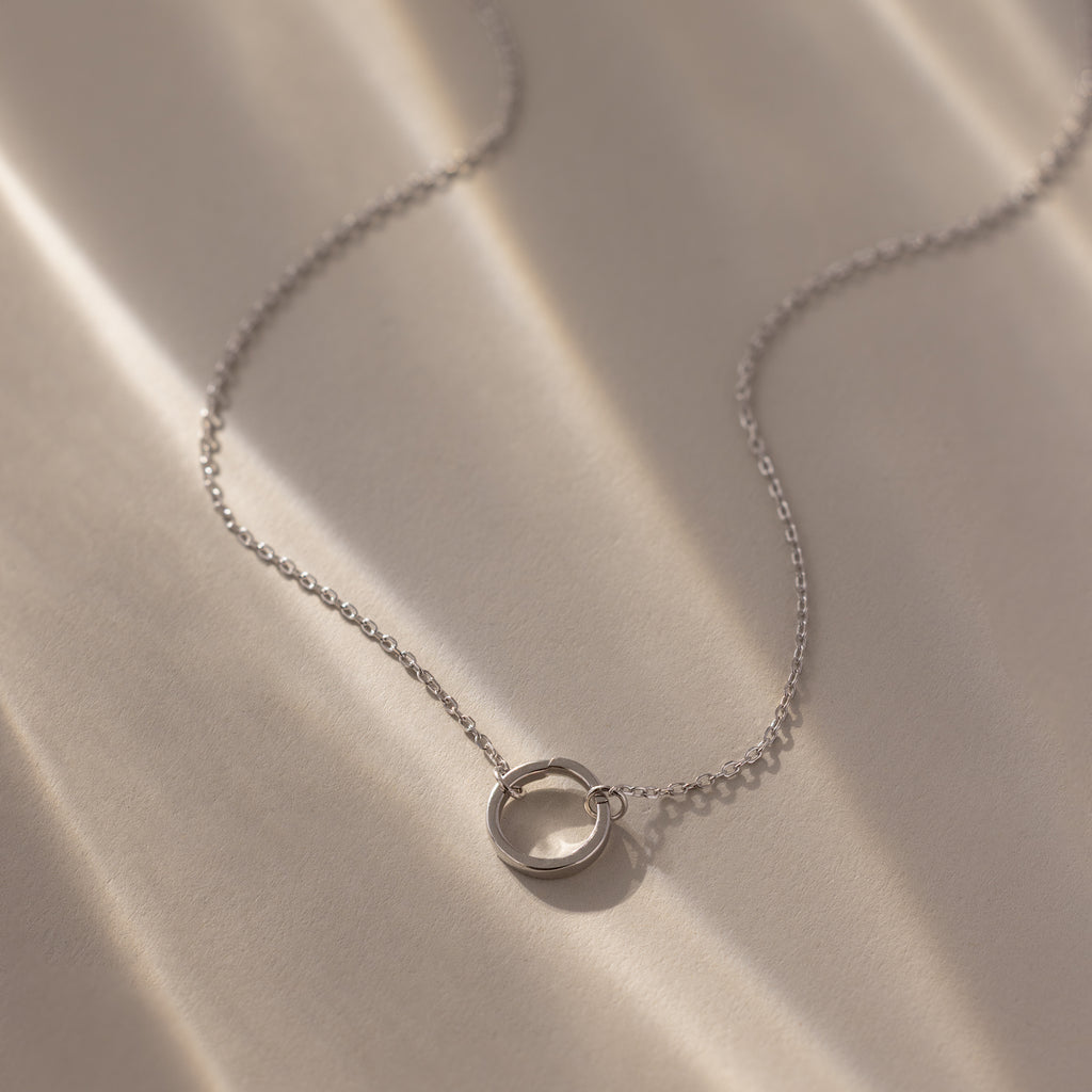 The Large Rolo Clasp Necklace features a delicate silver chain with a circular pendant and bold clasp, showcased on a softly lit beige surface.