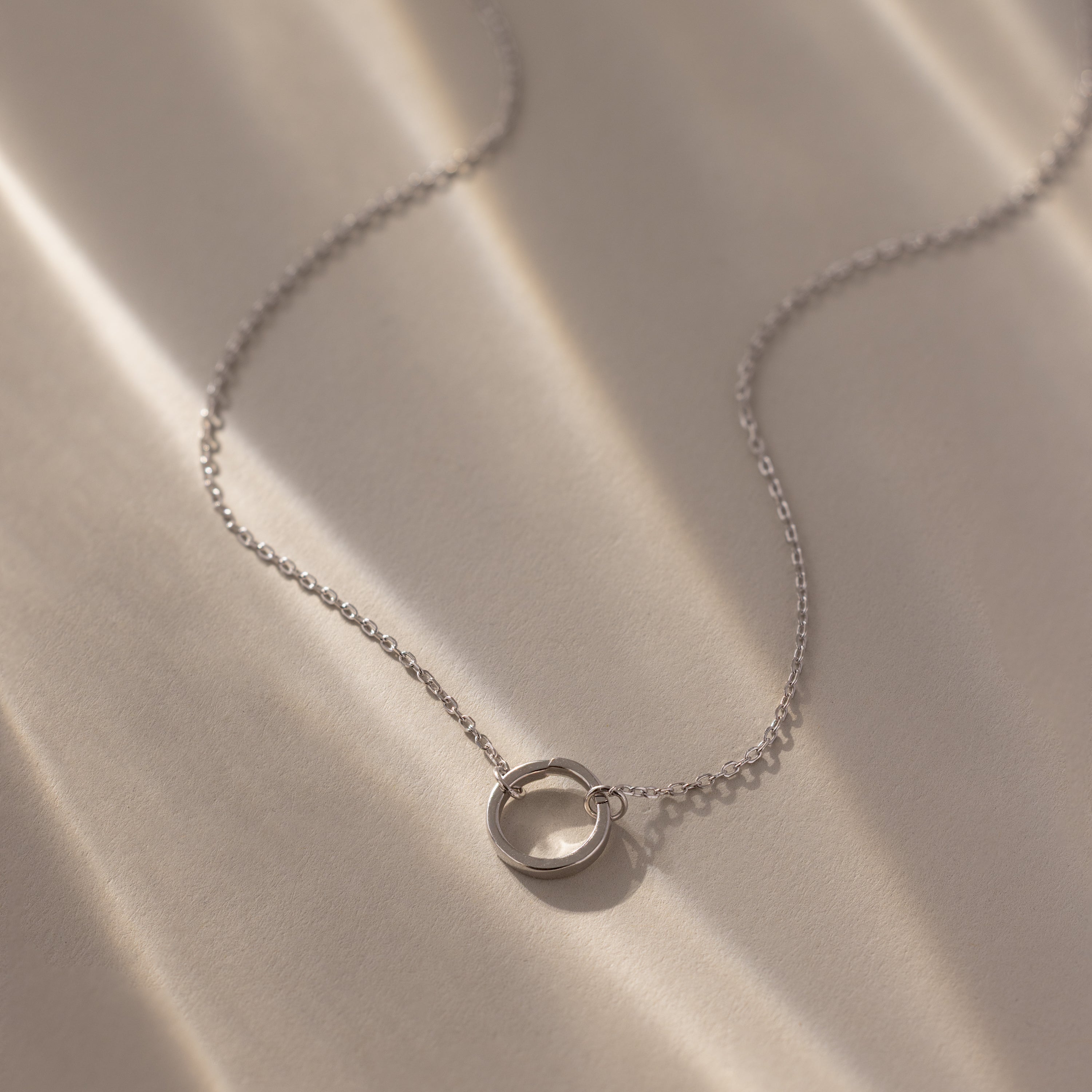 The Large Rolo Clasp Necklace features a delicate silver chain with a circular pendant and bold clasp, showcased on a softly lit beige surface.