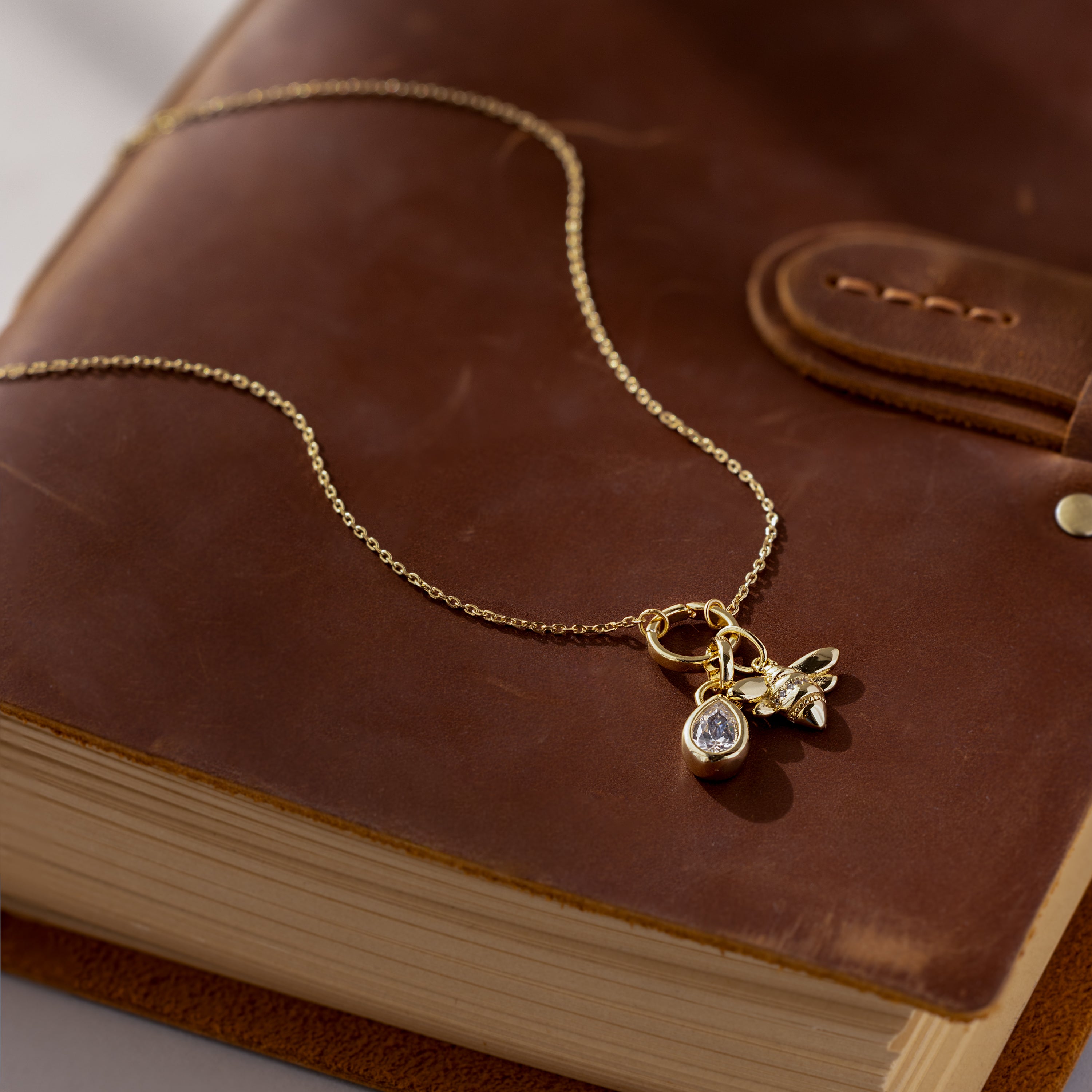 The Large Rolo Clasp Necklace in 18K Gold rests on a closed brown leather notebook, accented with a bee and gemstone charm—a stunning statement chain that’s perfect for layering.