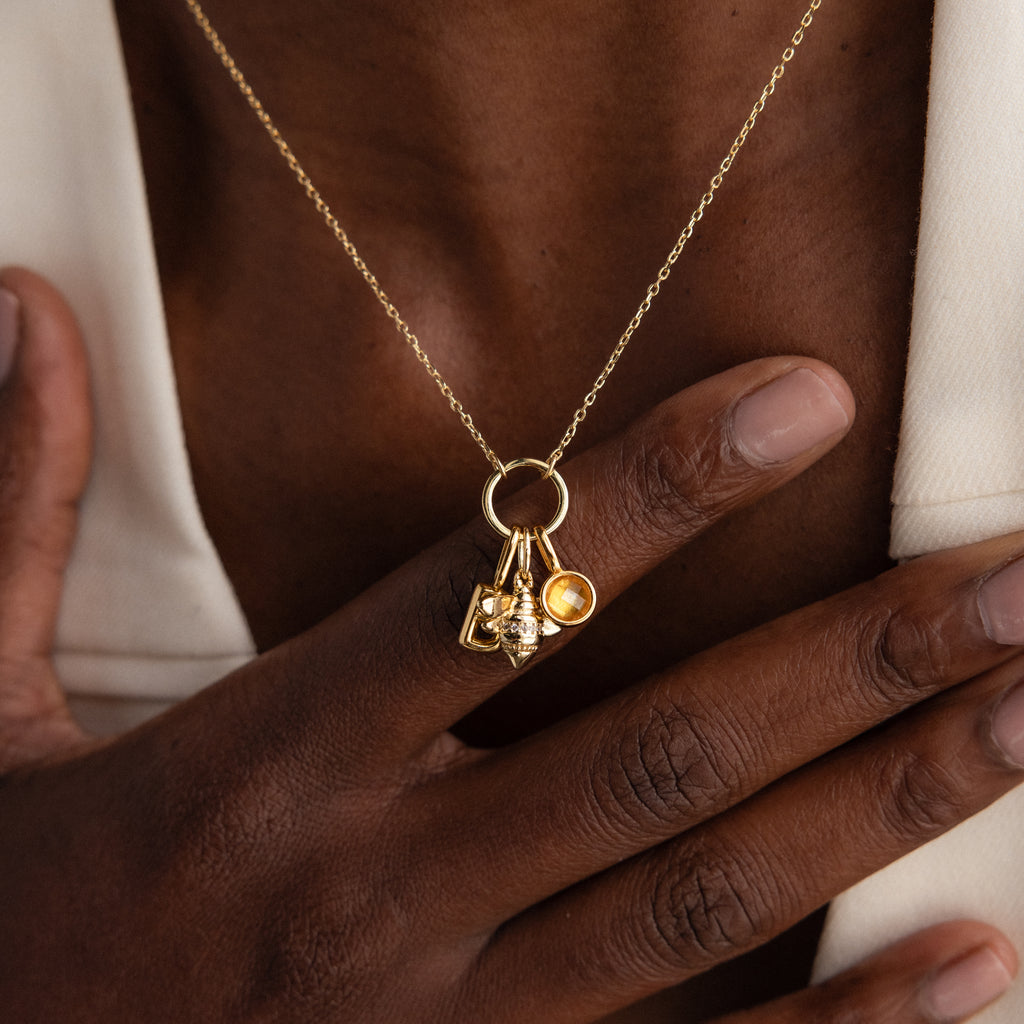 A hand touches the Large Rolo Clasp Necklace in 18K Gold, styled as a statement piece with multiple pendants over a cream blazer and bare chest.