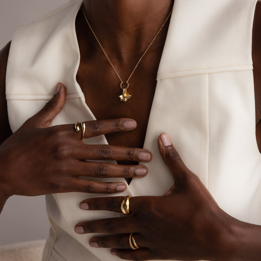 A person in a white vest, wearing gold rings and the Large Rolo Clasp Necklace in 18K Gold, rests their hands on their chest.