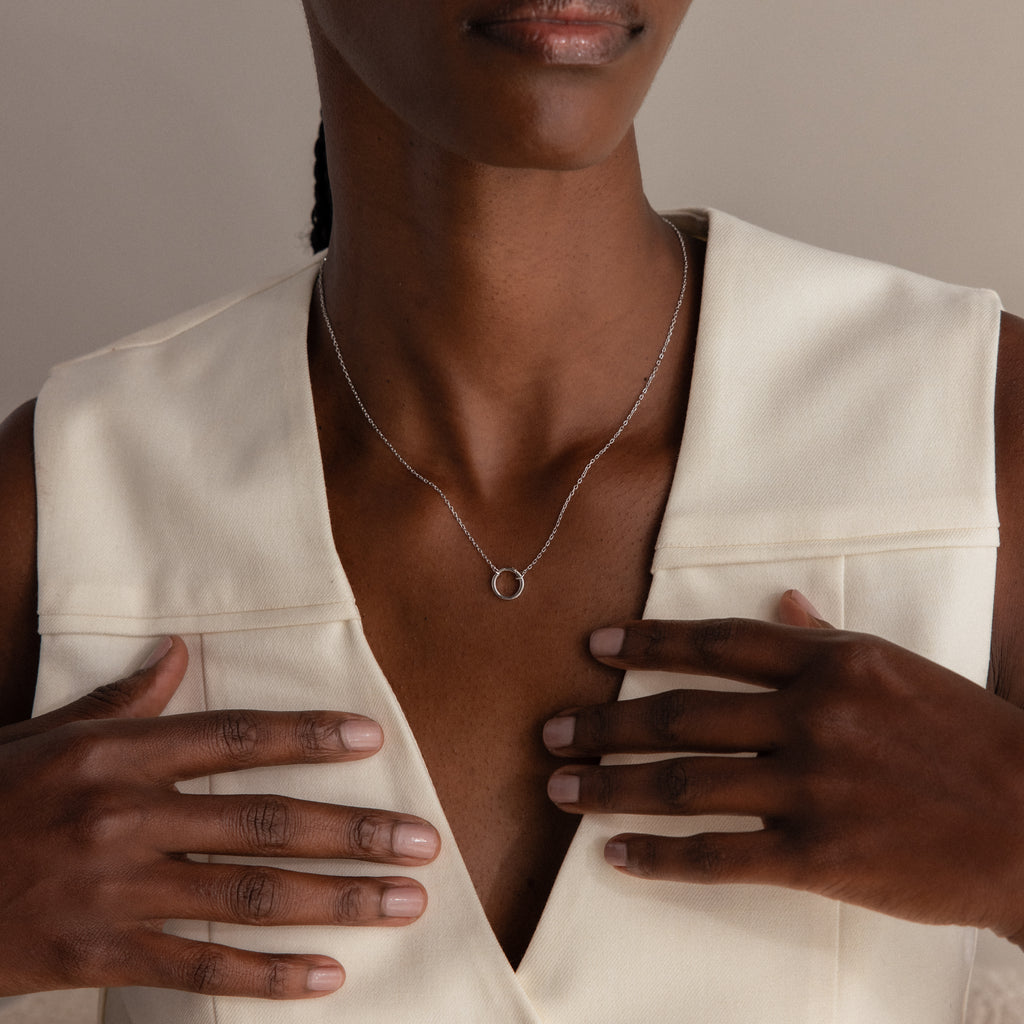 A woman in a cream sleeveless top wears the Large Rolo Clasp Necklace in Sterling Silver as a simple silver circle pendant, touching her chest—a versatile staple for any jewelry collection.