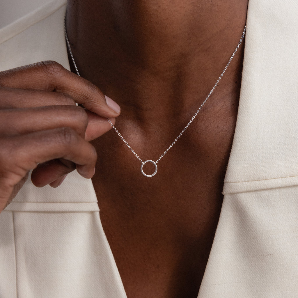 A person wearing the Large Rolo Clasp Necklace in Sterling Silver with a silver circle pendant and an off-white blazer, gently holding the pendant—a versatile and elegant staple for any wardrobe.