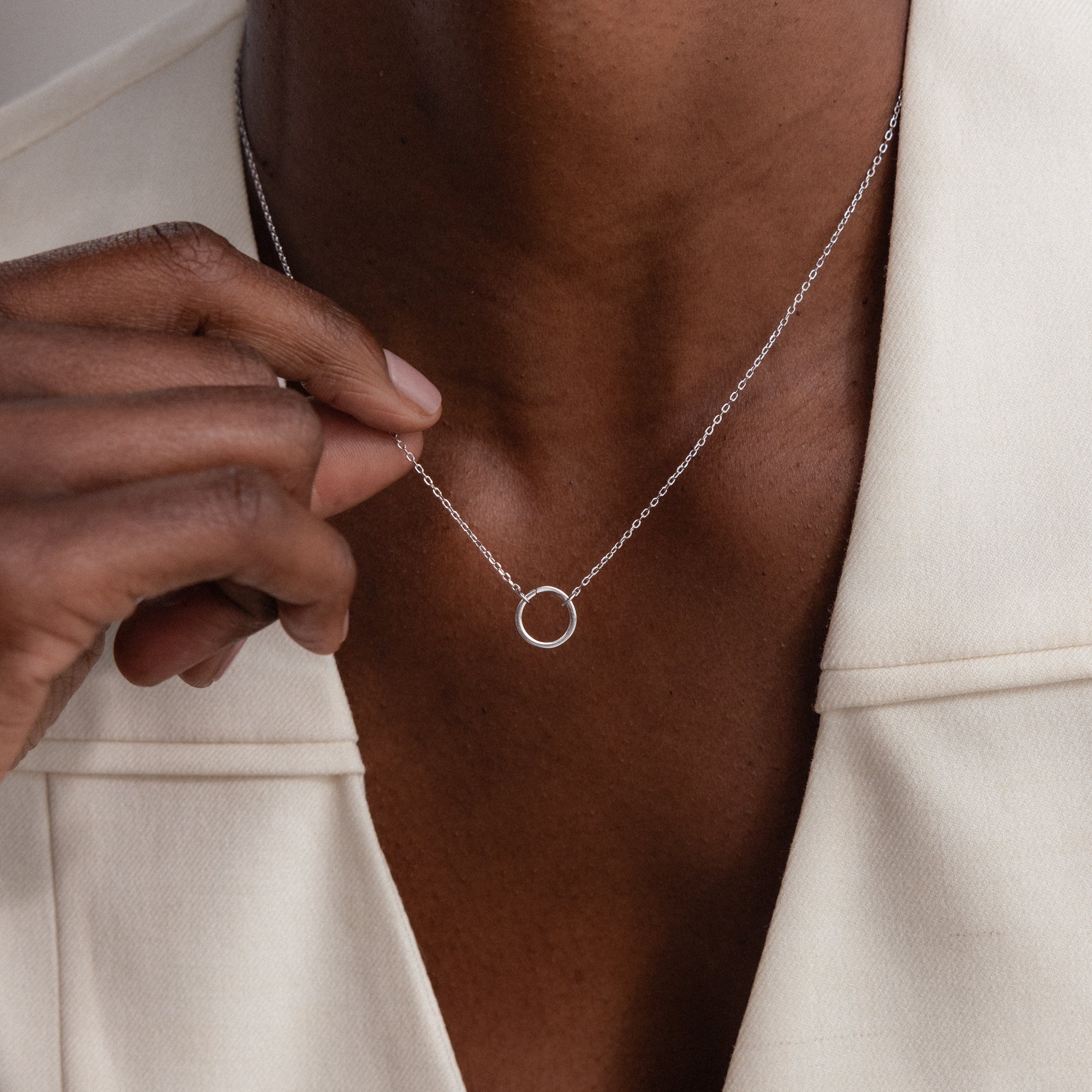 A person wearing the Large Rolo Clasp Necklace in Sterling Silver with a silver circle pendant and an off-white blazer, gently holding the pendant—a versatile and elegant staple for any wardrobe.