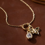 Modern essential: The Large Rolo Clasp Necklace showcases a bold statement chain with a gold teardrop crystal and bee charm, styled on a brown surface.