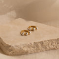 The Triple Pearl Huggies—gold hoops with three white pearls—sit on a textured beige stone, offering a modern, minimalist touch for lovers of personalized jewelry.