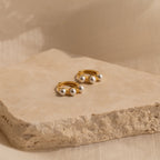 The Triple Pearl Huggies—gold hoops with three white pearls—sit on a textured beige stone, offering a modern, minimalist touch for lovers of personalized jewelry.