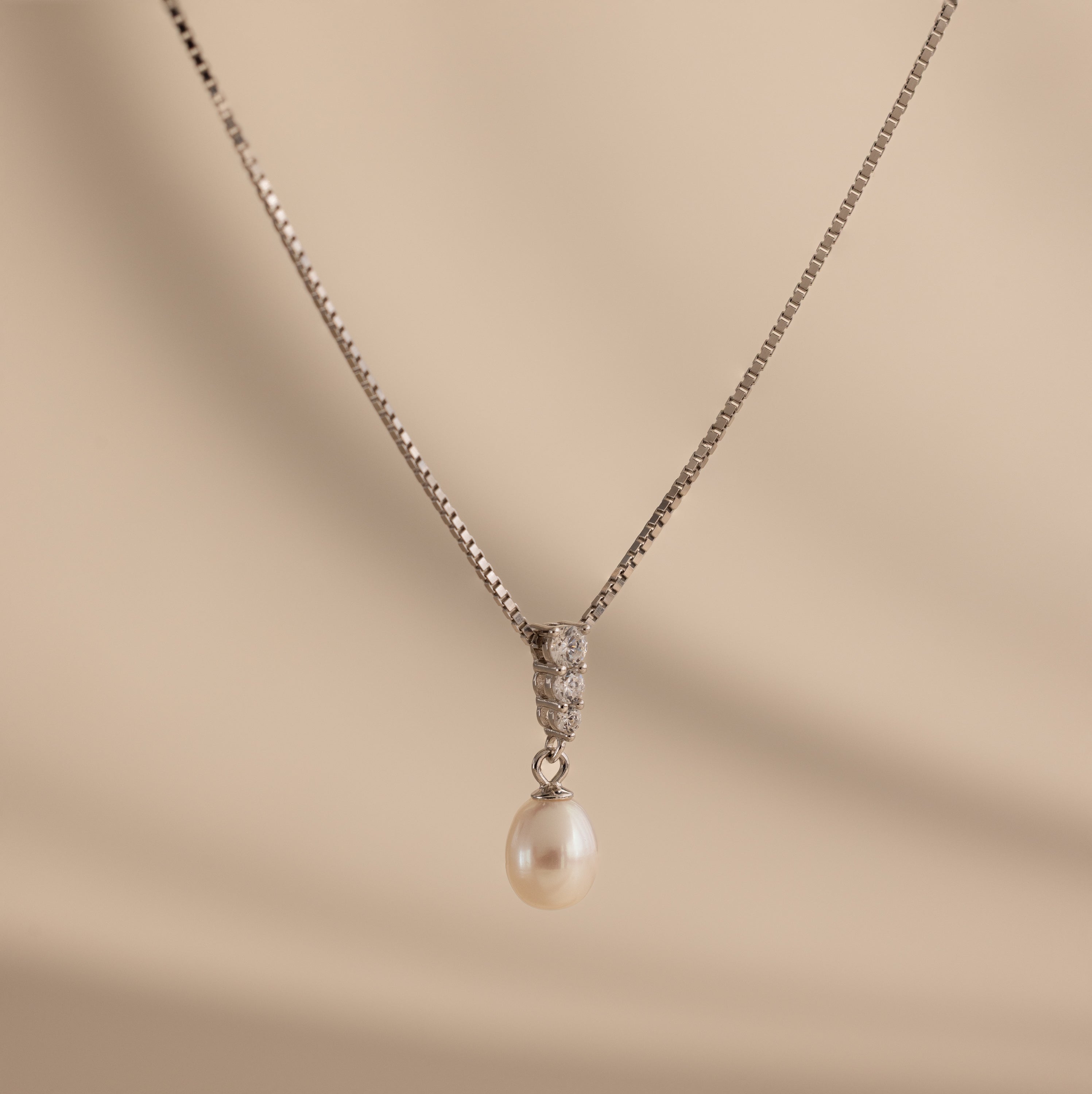 Diamond Pearl Droplet Necklace in Sterling Silver features a diamond-studded drop pendant with a single white pearl, showcased against a beige background.