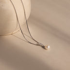 The Diamond Pearl Droplet Necklace in Sterling Silver, featuring a single pearl in a delicate drop design, is displayed on a light beige surface next to a round vase.