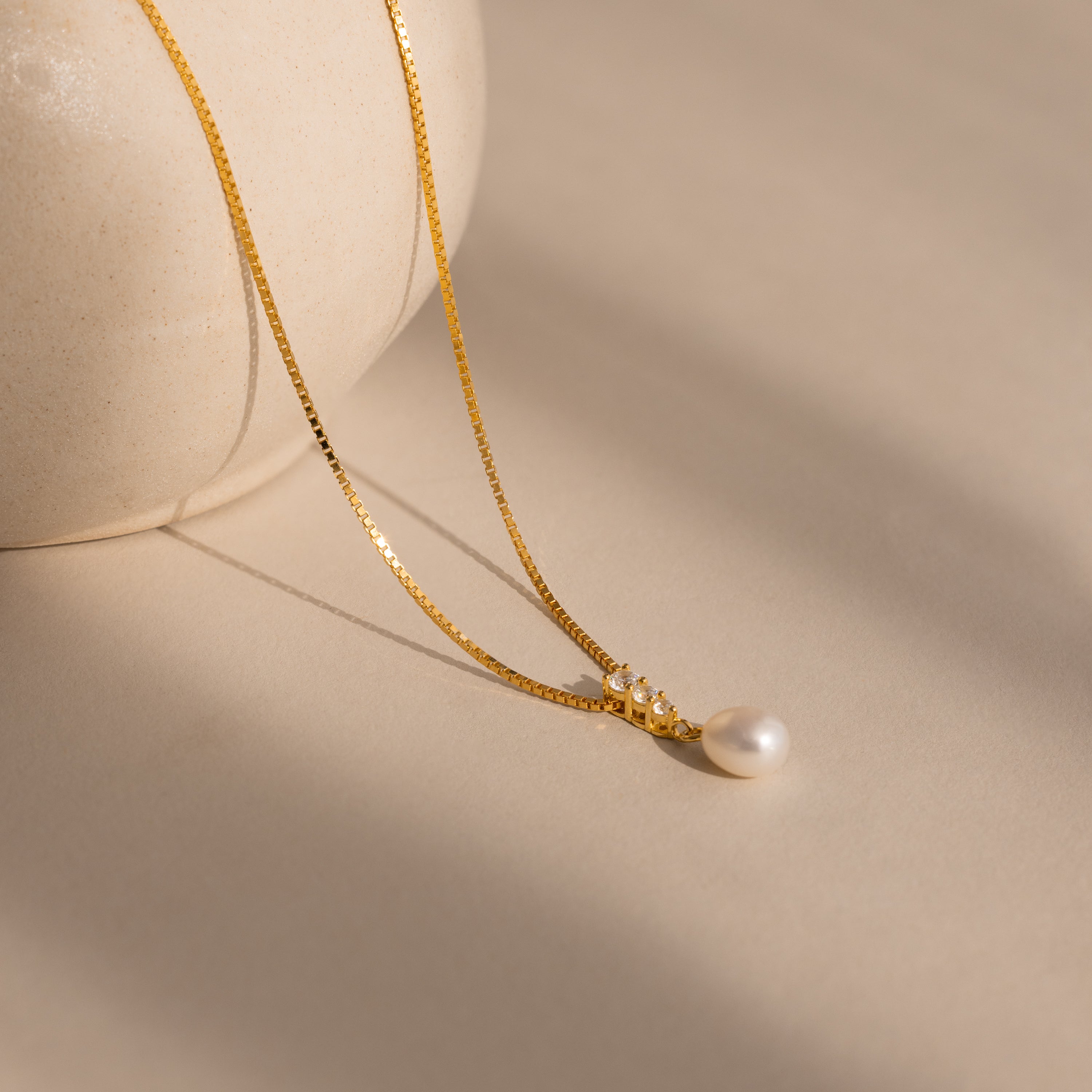 The Diamond Pearl Droplet Necklace in 18K Gold features a gold chain and pearl pendant, elegantly displayed on a neutral surface beside a round, light-colored vase.