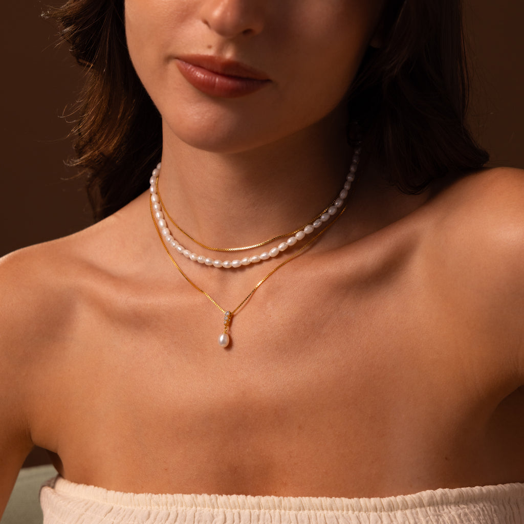 A woman wearing the Diamond Pearl Droplet Necklace layered with gold necklaces and an off-shoulder top, set against a soft brown background.
