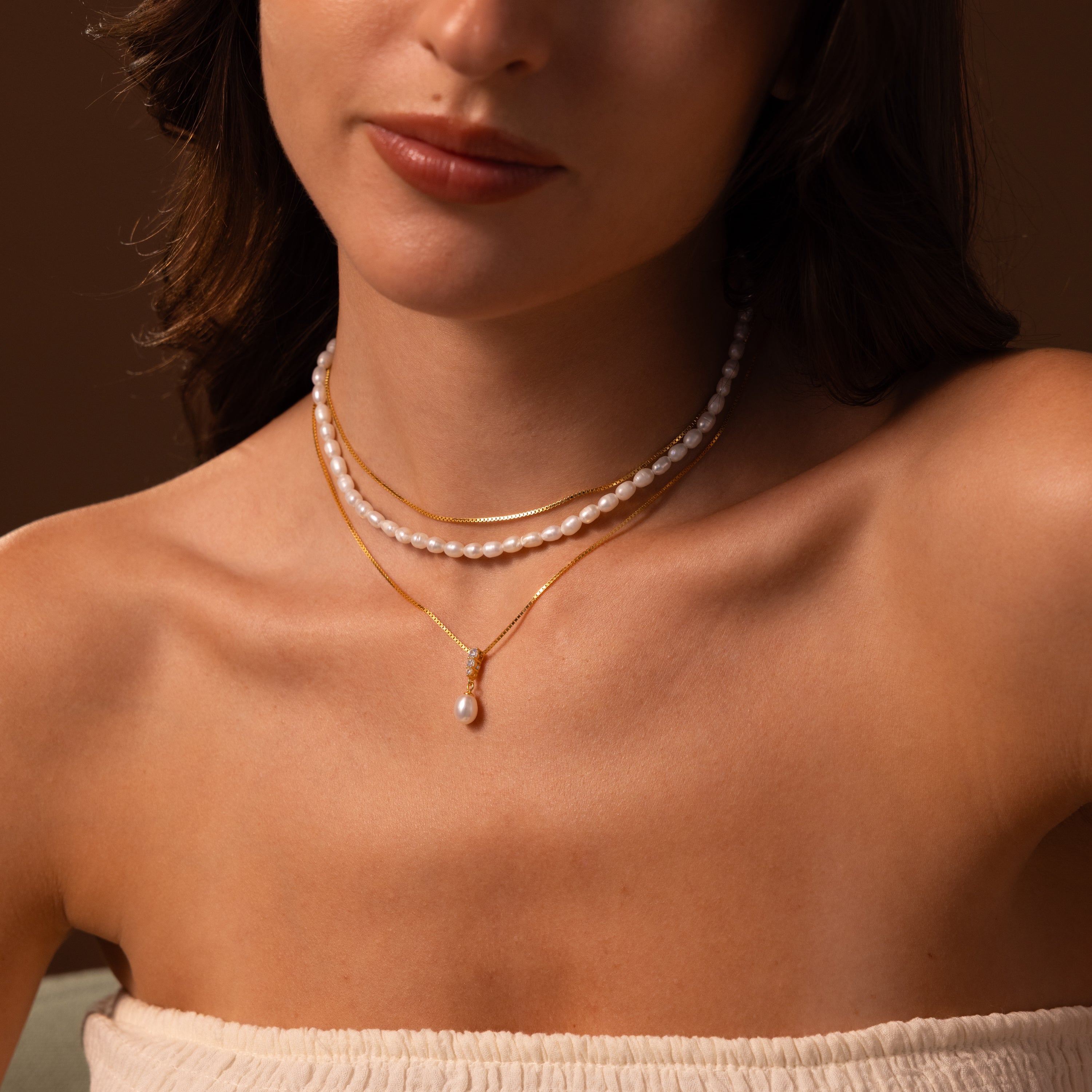A woman wearing the Diamond Pearl Droplet Necklace layered with gold necklaces and an off-shoulder top, set against a soft brown background.