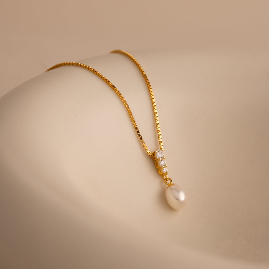 Diamond Pearl Droplet Necklace in 18K Gold, featuring a gold chain with a pearl pendant and delicate gems, displayed on a smooth beige surface.