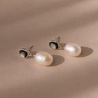 The Midnight Pearl Drop Earrings in Sterling Silver, featuring elegant silver and black accents, rest on a beige surface under soft lighting.
