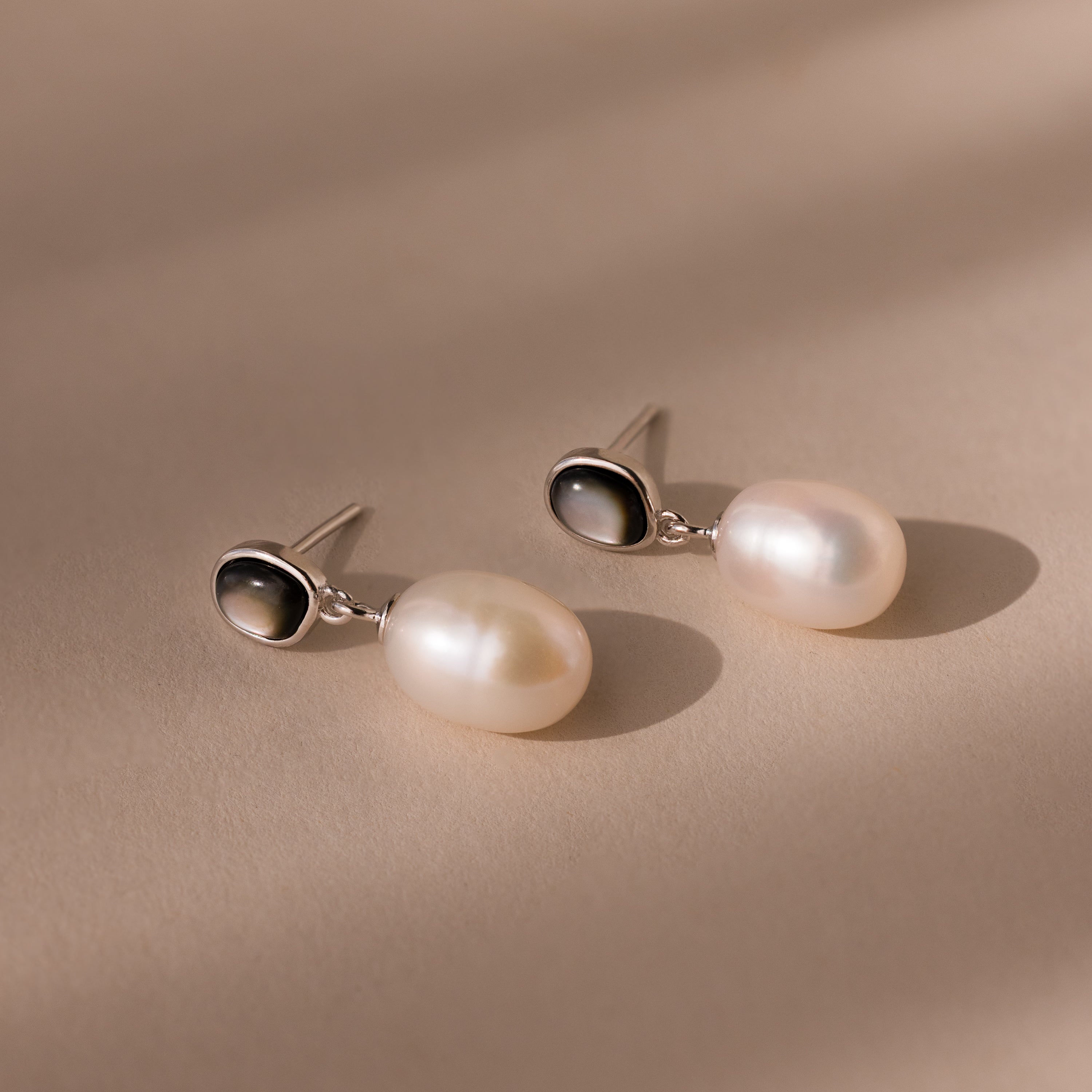 The Midnight Pearl Drop Earrings in Sterling Silver, featuring elegant silver and black accents, rest on a beige surface under soft lighting.
