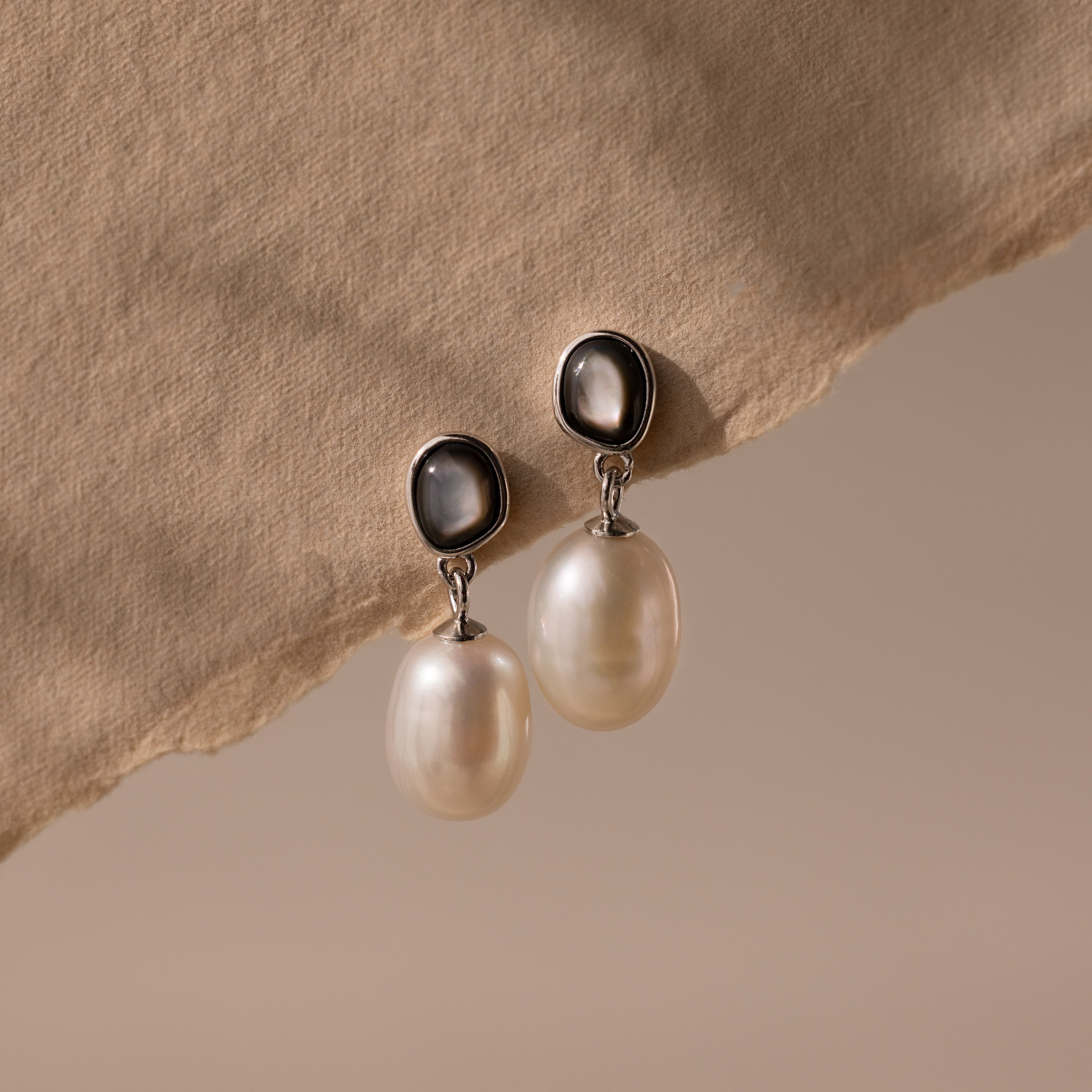 The Midnight Pearl Drop Earrings in Sterling Silver feature dark oval studs with subtle black accents, elegantly showcased on a beige fabric—an exquisite piece for any timeless jewelry collection.