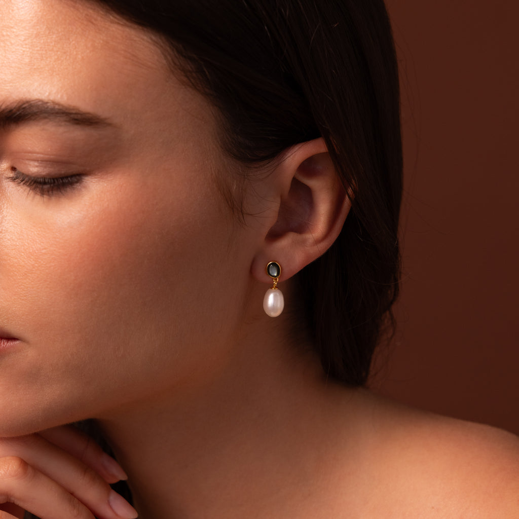 A woman in profile touches her chin, showcasing the Midnight Pearl Drop Earrings in 18K Gold—timeless jewelry with elegant pearl drops and subtle black accents—against a warm brown background.