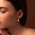 A woman in profile touches her chin, showcasing the Midnight Pearl Drop Earrings in 18K Gold—timeless jewelry with elegant pearl drops and subtle black accents—against a warm brown background.