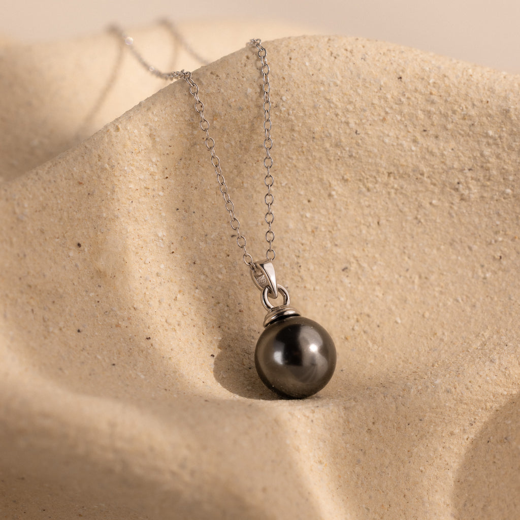 The Midnight Pearl Necklace showcases a dark round pearl pendant on a silver chain, elegantly displayed on textured beige fabric—a timeless jewelry piece.