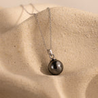 The Midnight Pearl Necklace showcases a dark round pearl pendant on a silver chain, elegantly displayed on textured beige fabric—a timeless jewelry piece.