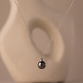 The Midnight Pearl Necklace in Sterling Silver features a silver chain with a black pearl pendant, showcased on a neutral abstract background—an exquisite piece of elegant jewelry.