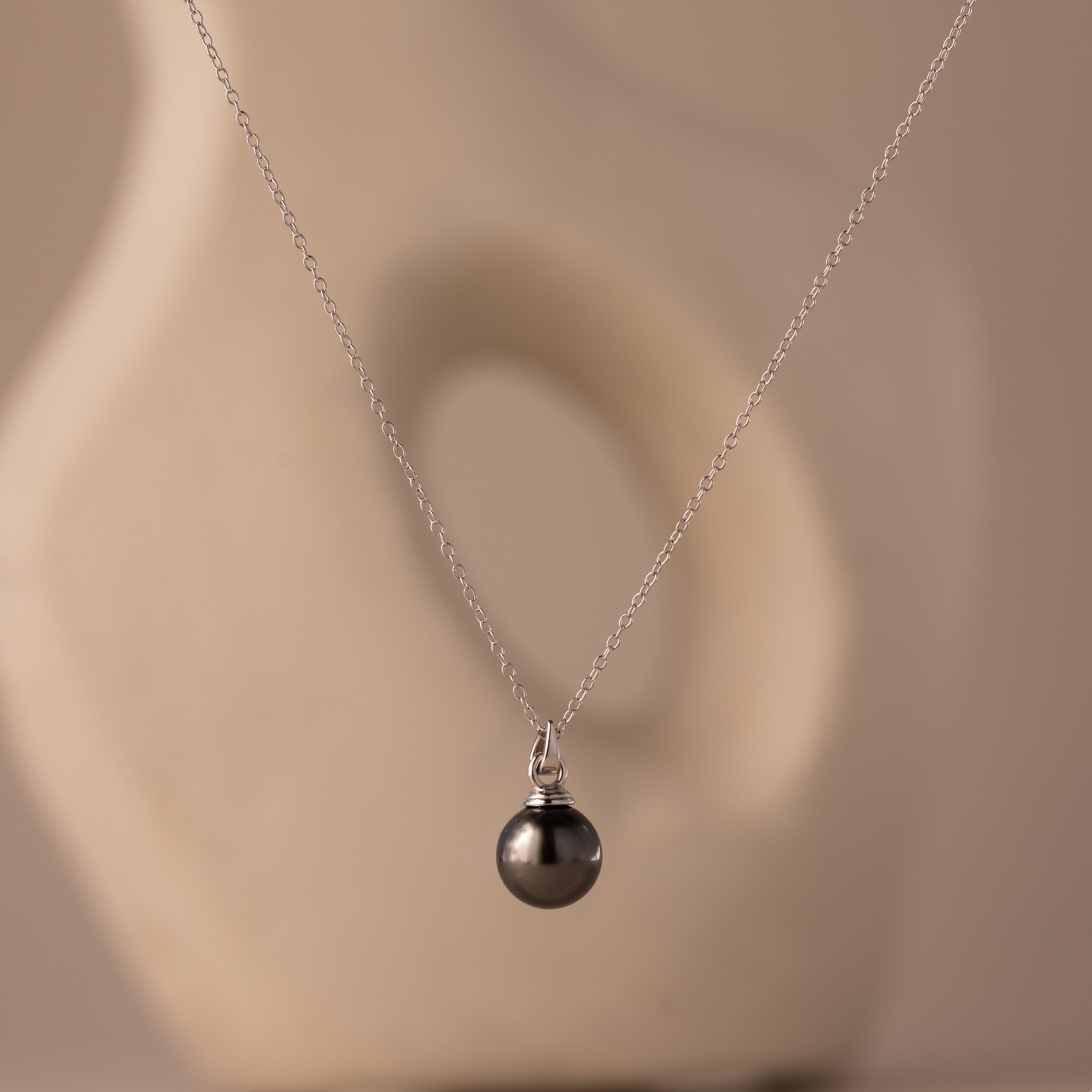 The Midnight Pearl Necklace in Sterling Silver features a silver chain with a black pearl pendant, showcased on a neutral abstract background—an exquisite piece of elegant jewelry.